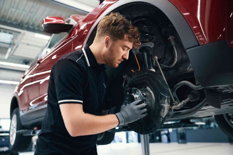 BMW Service 5+ 8+ 4 Ein Mechaniker mit Handschuhen überprüft und repariert die Bremsen eines roten Autos, das in einer gut beleuchteten Werkstatt auf einer Hebebühne steht.