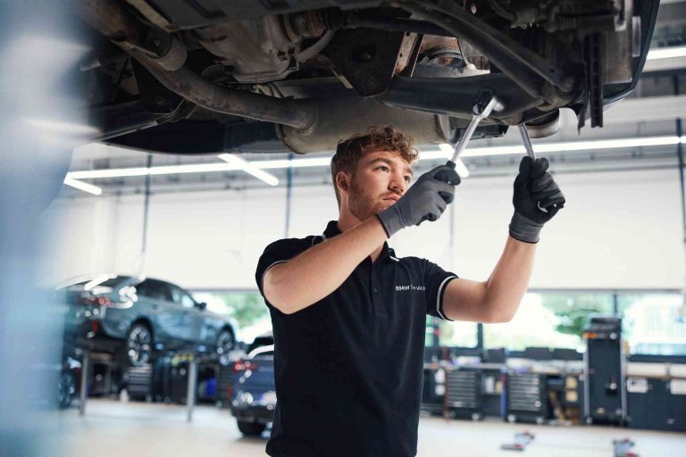 BMW Service 5+ 8+ 3 In einer hellen, modernen Autowerkstatt arbeitet ein Mechaniker mit Handschuhen und einem Schraubenschlüssel an der Unterseite eines angehobenen Fahrzeugs.