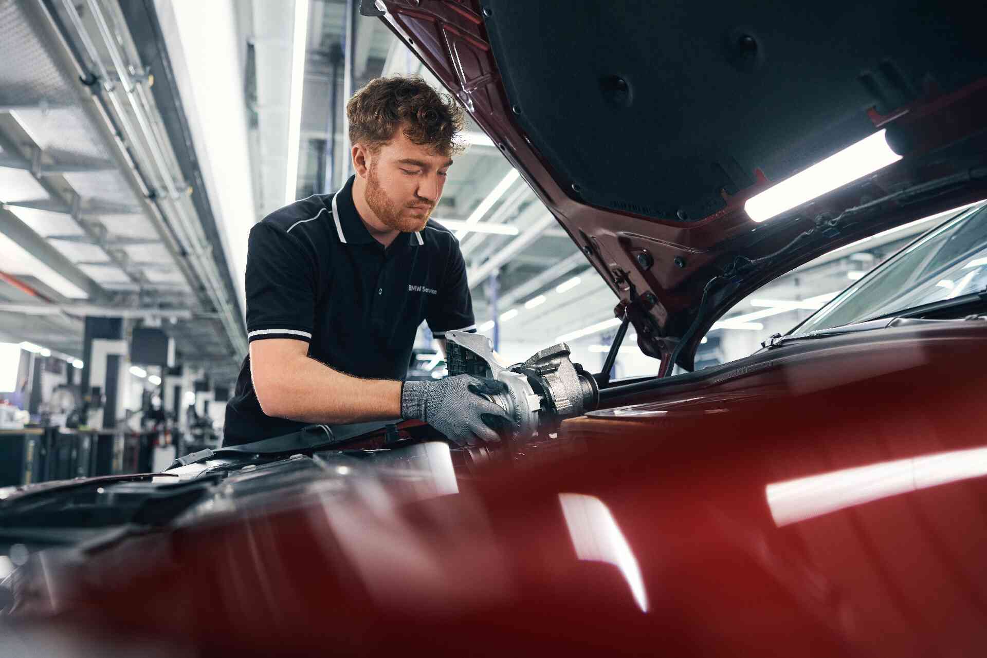 BMW Service 5+ 8+ 2 Ein Mechaniker mit Handschuhen arbeitet in einer gut beleuchteten Autowerkstatt unter der Motorhaube eines roten Autos und konzentriert sich auf ein Autoteil, während er von Werkzeugen und Geräten umgeben ist.
