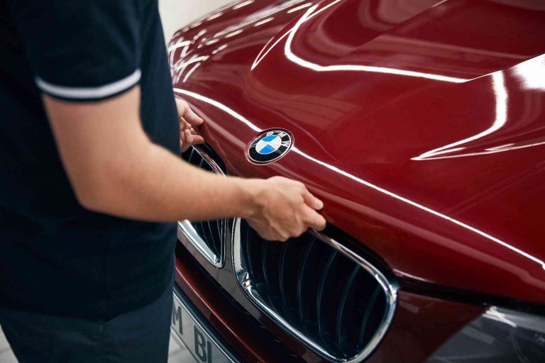 BMW Service 5+ 8+ 1 Eine Person steht vor einem leuchtend roten BMW und ist gerade dabei, die Motorhaube zu öffnen. Zu sehen sind nur ihre Arme und ihr Oberkörper. Das BMW-Logo ist deutlich auf der Motorhaube zu erkennen.