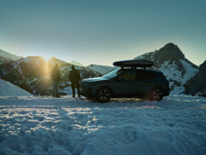 ein dunkler BMW iX mit einer BMW Dachbox steht auf einem zugeschneiten Berg und 2 Personen stehen vor dem Auto