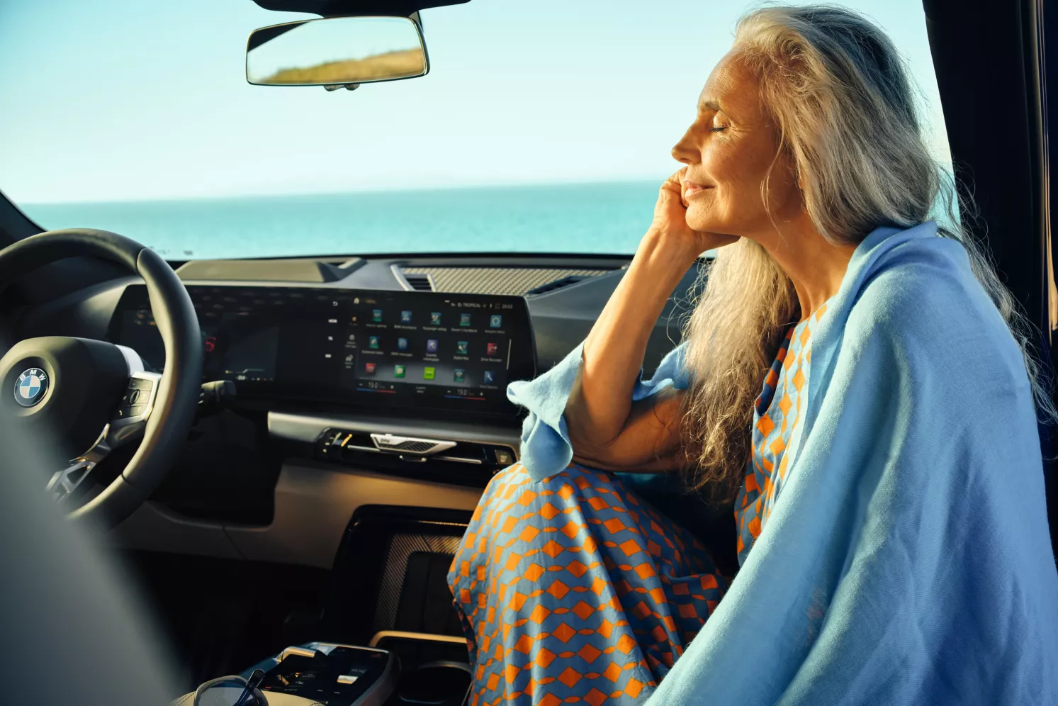 Eine ältere Frau mit langen grauen Haaren sitzt in einem Auto am Meer und lächelt friedlich. Sie trägt ein blau-orangefarbenes Kleid mit einem blauen Schal, und das Sonnenlicht fällt durch das Fenster ein. Das Armaturenbrett des BMW ist zu sehen.