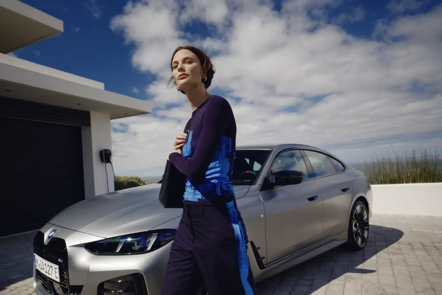 Eine Frau mit kurzen roten Haaren steht selbstbewusst vor einem silbernen BMW, der vor einem modernen Haus unter einem teilweise bewölkten Himmel geparkt ist.