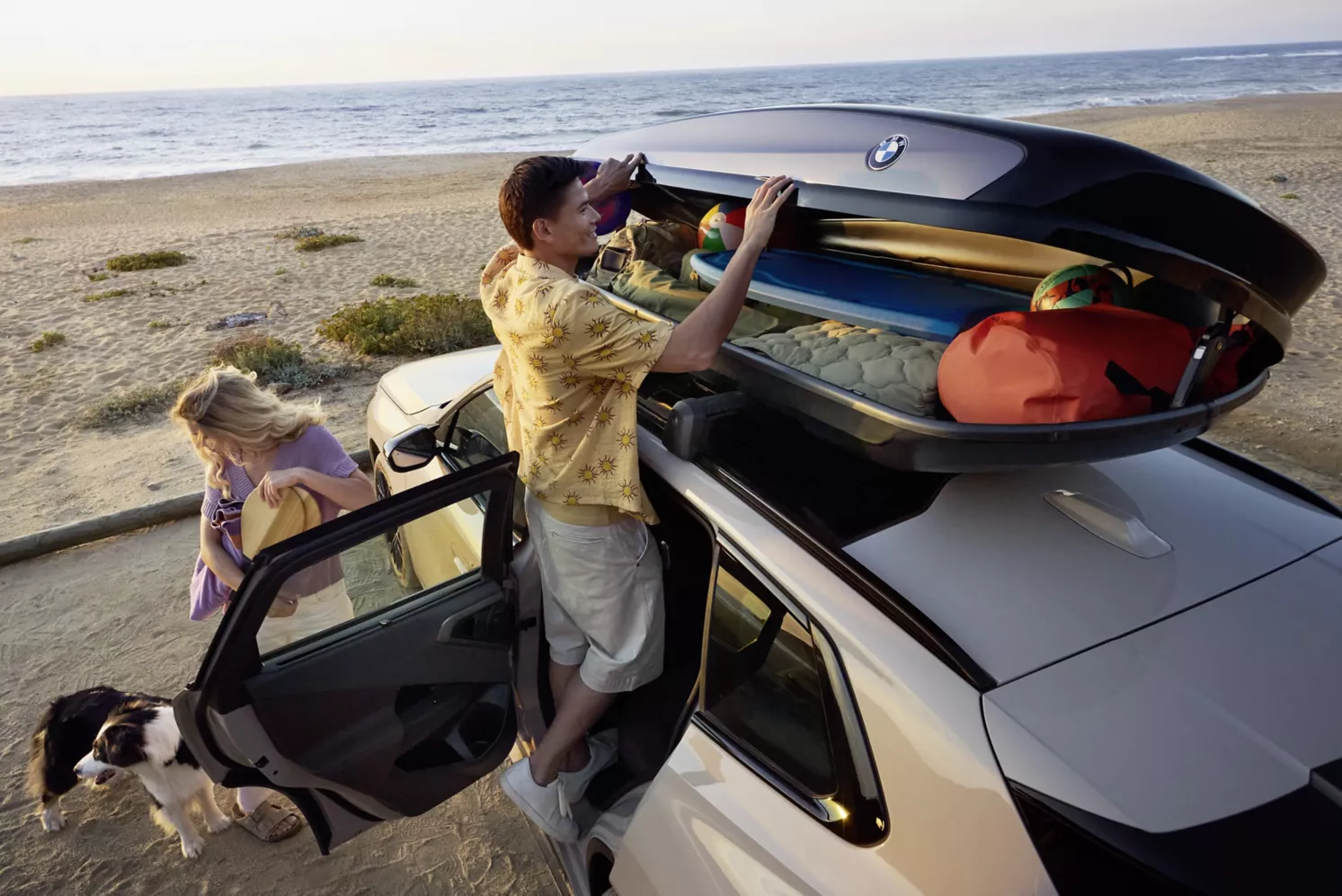 Ein Mann lädt Strandutensilien in eine Dachbox eines weißen Geländewagens, der am Meer geparkt ist, während eine Frau mit einem Hund in der Nähe steht. Im Hintergrund geht die Sonne über dem Strand unter.