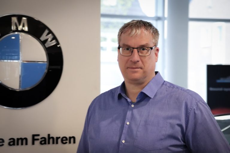 Ein Mann mit Brille und blauem Hemd steht in einem Haus neben einem BMW-Logo an der Wand. Der Hintergrund zeigt große Fenster und einen Teil eines Schildes mit deutschem Text.