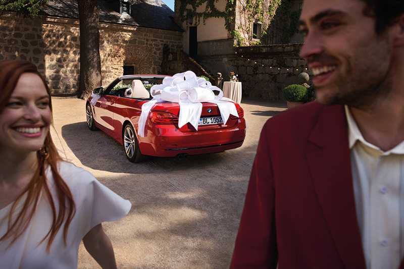Im Vordergrund steht ein lächelndes Paar, während hinter ihnen ein rotes Cabrio mit einer großen weißen Schleife geparkt ist, was auf einen festlichen Anlass wie eine Hochzeit oder ein besonderes Ereignis hindeutet.