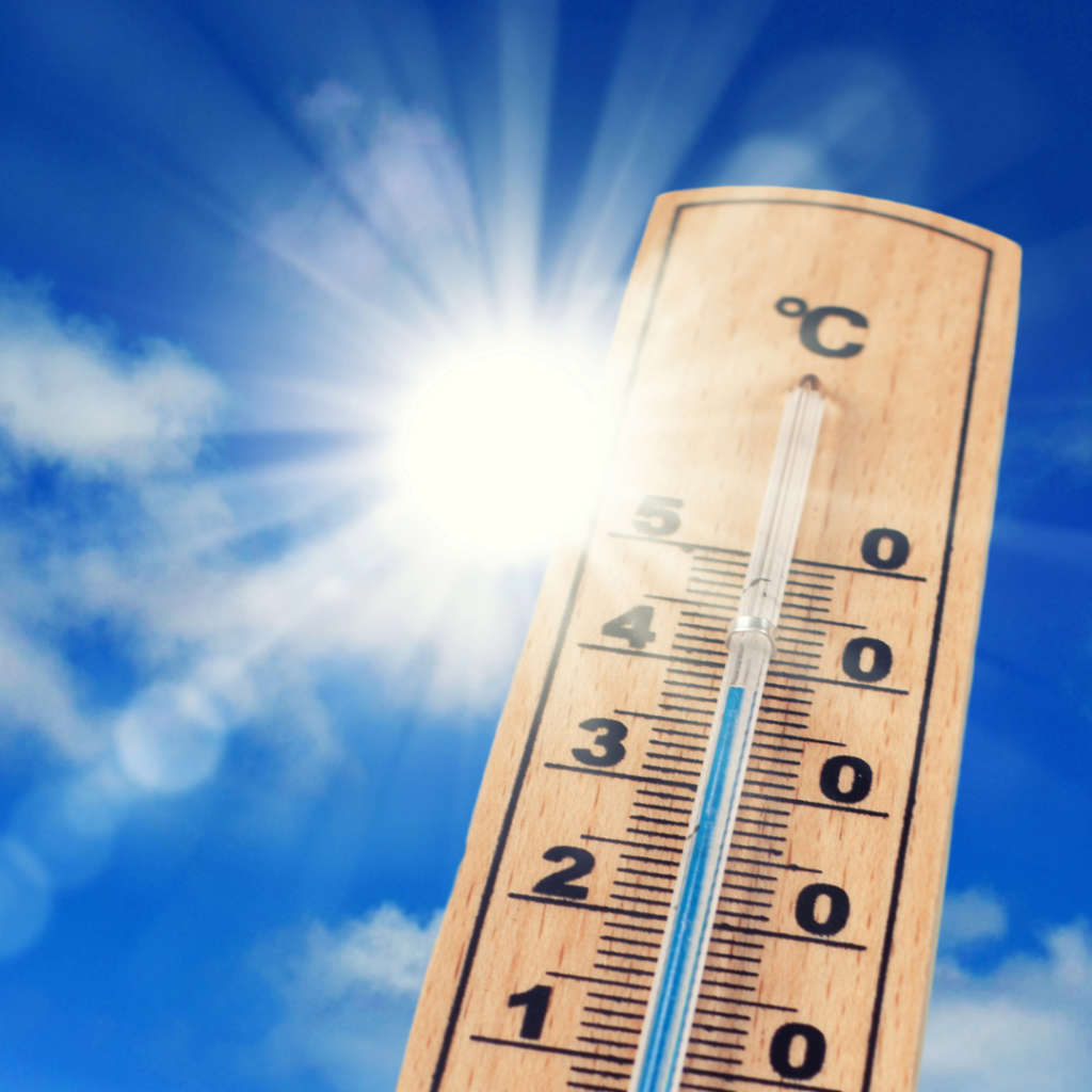 Eine Nahaufnahme eines Holzthermometers zeigt hohe Temperaturen in Celsius, im Hintergrund sind die strahlende Sonne und ein blauer Himmel mit einigen Wolken zu sehen, was auf heißes Wetter hindeutet.