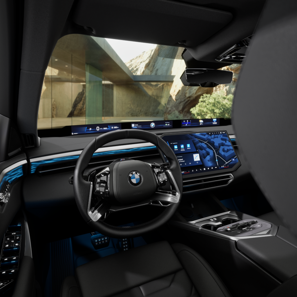 Moderner BMW-Innenraum mit digitalem Armaturenbrett und Lenkrad, ausgestattet mit mehreren Bildschirmen und Ambientebeleuchtung. Durch die großen Fenster ist draußen ein modernes Gebäude und eine felsige Landschaft zu sehen.