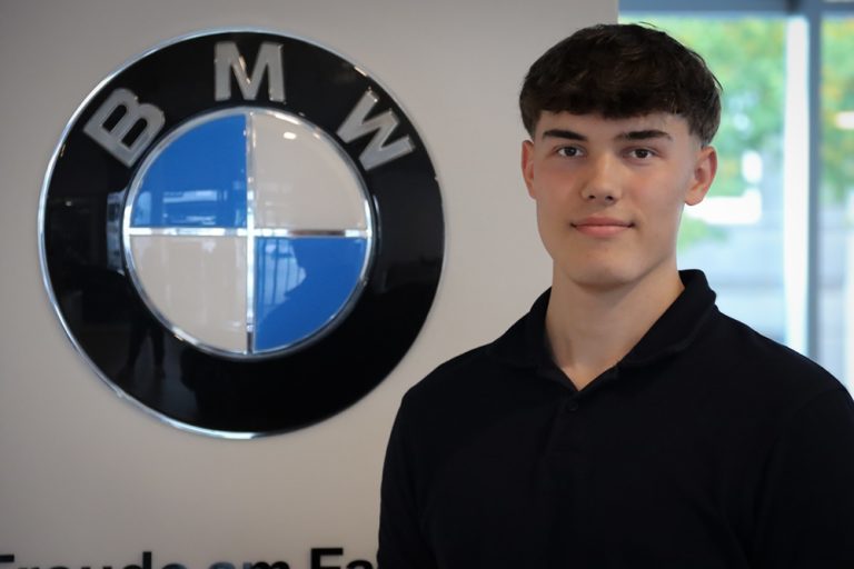 Ein junger Mann in einem dunklen Polohemd steht neben einem großen BMW-Logo an einer Wand. Im Hintergrund sind ein verschwommenes Fenster und Grünflächen zu sehen.