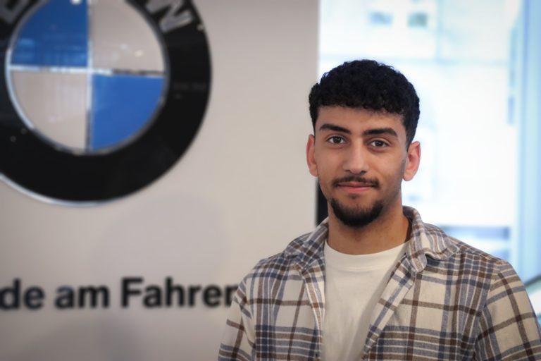 Ein junger Mann mit kurzen lockigen Haaren und einem Bart, der ein kariertes Hemd trägt, steht in einem Innenraum vor einem BMW-Logo und einem verschwommenen Schild.