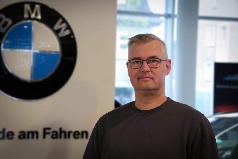 Ein Mann mit kurzen grauen Haaren und Brille steht in einem Haus vor einem großen BMW-Logo und einem Teil des Textes auf einem Schild, mit verschwommenen Fenstern und Autos im Hintergrund.