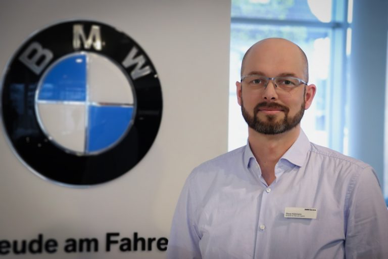 Ein Mann mit Brille, Bart und hellblauem Hemd steht in einem Innenraum neben einem BMW-Logo-Display. Er trägt ein Namensschild, und die Umgebung scheint ein BMW-Ausstellungsraum oder ein Autohaus zu sein.